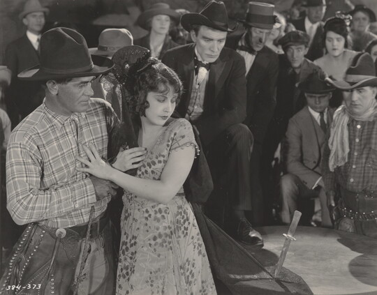 A black-and-white photograph of a White cowboy comforting a White woman whose veil is pinned to a table with a knife, as a crowd looks on behind her.