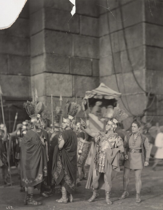 A black-and-white photograph of a group of people dressed as ancient Romans in front of a large stone building.