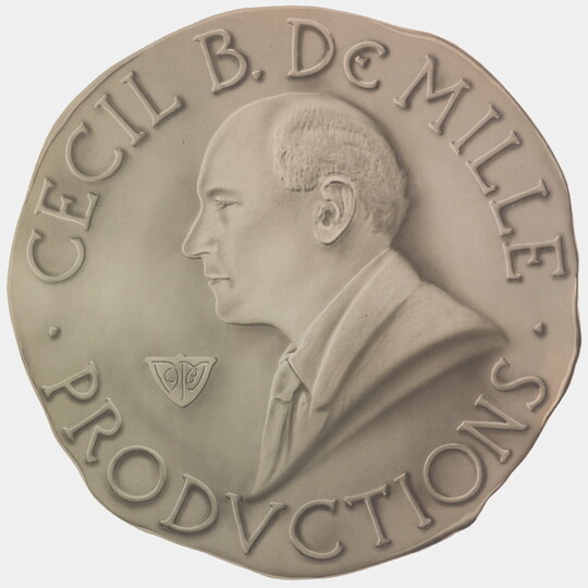 A sepia-toned photograph of a coin with a profile of a balding White man and the words "Cecil B. DeMille Productions" around the perimeter.