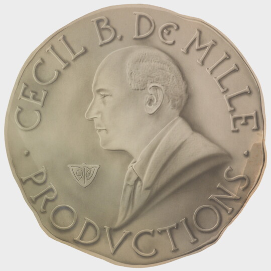 A sepia-toned photograph of a coin with a profile of a balding White man and the words "Cecil B. DeMille Productions" around the perimeter.
