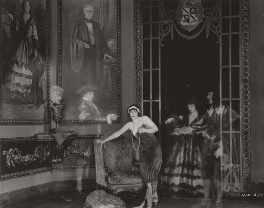 A black-and-white photograph of a White woman standing next to a loveseat in a lavish room as four transparent people in historical costumes point to her.