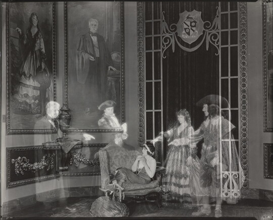 A black-and-white photograph of a White woman huddled on a loveseat in a lavish room as four transparent people in historical costumes point to her.