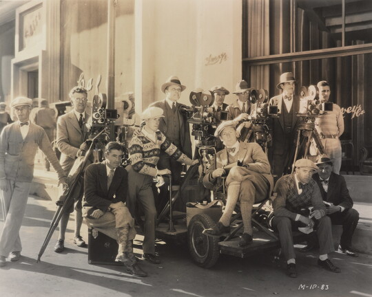 A black-and-white photograph of a group of men gathered outside of a building with film equipment.
