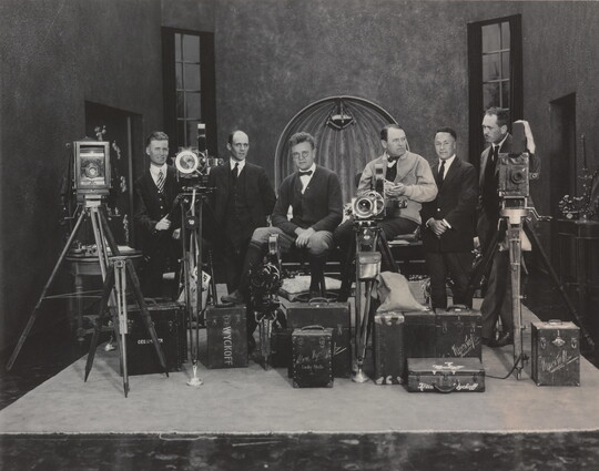A black-and-white photograph of a group of White men in suits on a movie set posed with multiple cameras and lighting equipment.
