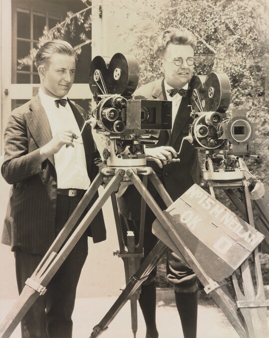 A black-and-white photograph of two White men standing next to each other behind two large video cameras.