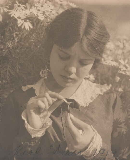 A black-and-white photograph of a young White woman picking the last petal off of a flower she holds in her hand.