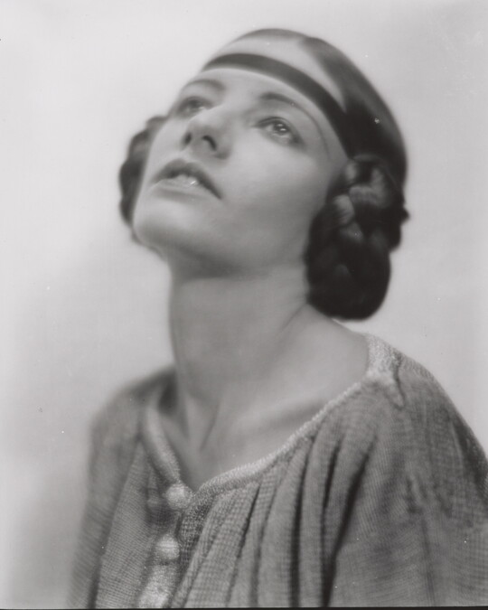A black-and-white portrait of a White woman, head angled up, with her hair braided into buns on the sides of her head.