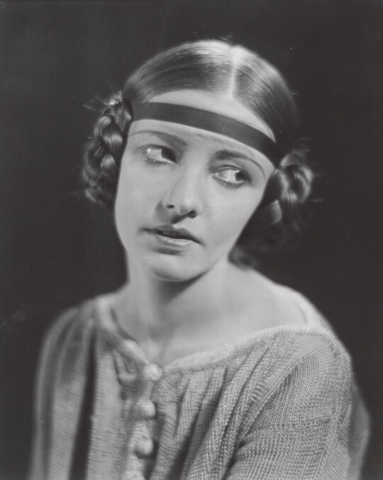 A black-and-white portrait of a White woman, eyes looking to the side, with her hair braided into buns on the sides of her head.