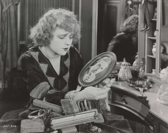A black-and-white photograph of a White woman, seated at a vanity, gazing down at a photograph of a White man in an oval frame.
