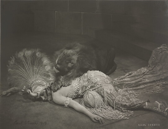 A black-and-white photograph of an elaborately dressed White woman lying on the ground with a snarling lion lying over her back.