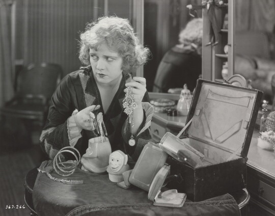 A black-and-white photograph of a White woman seated at a vanity, holding two handfuls of jewelry and looking off to the side.
