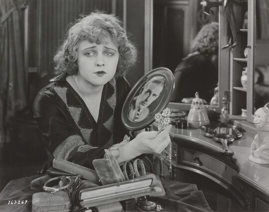 A black-and-white photograph of a White woman sitting at a vanity holding a brooch and a photograph of a White man as she gazes into the distance.