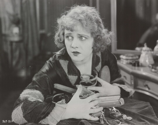 A black-and-white photograph of a White woman seated at a vanity clutching a variety of objects and jewelry to her chest.