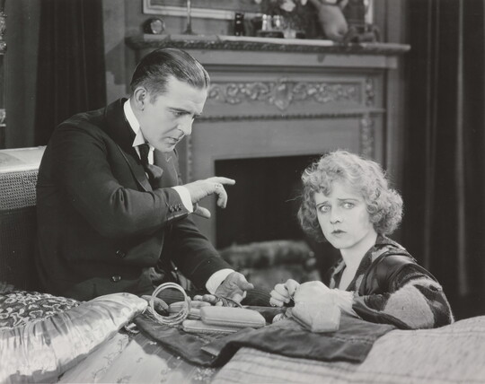 A black-and-white photograph of a White man gesturing toward a seated White woman who is looking toward the viewer.