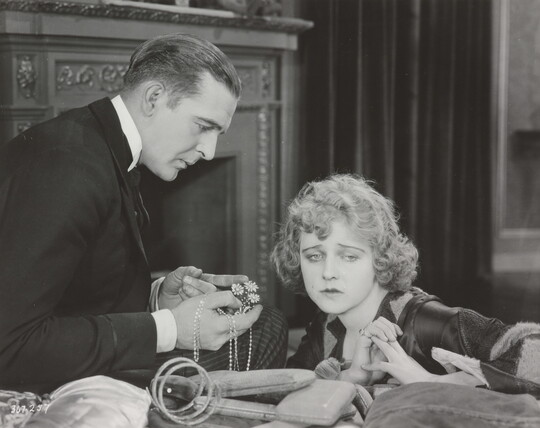 A black-and-white photograph of a White man presenting a White woman with jewelry as she sadly gazes into the distance.