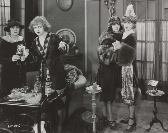 A black-and-white photograph of four White women standing in a room looking at something in the distance, two of the women holding each other in surprise.