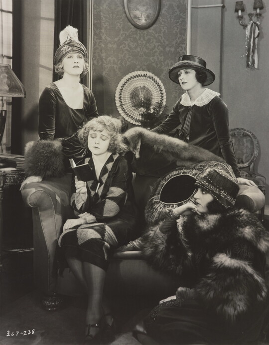 A black-and-white photograph of four elaborately-dressed White women, two standing, one reading on a sofa, the other seated on the floor.