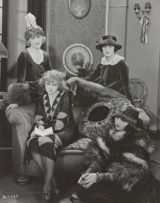 A black-and-white photograph of four elaborately-dressed White women, two standing, one on a sofa with a book in her lap, the other seated on the floor.