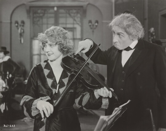 A black-and-white photograph of a White woman playing a violin and an older White man next to her with his finger violin's chinrest.