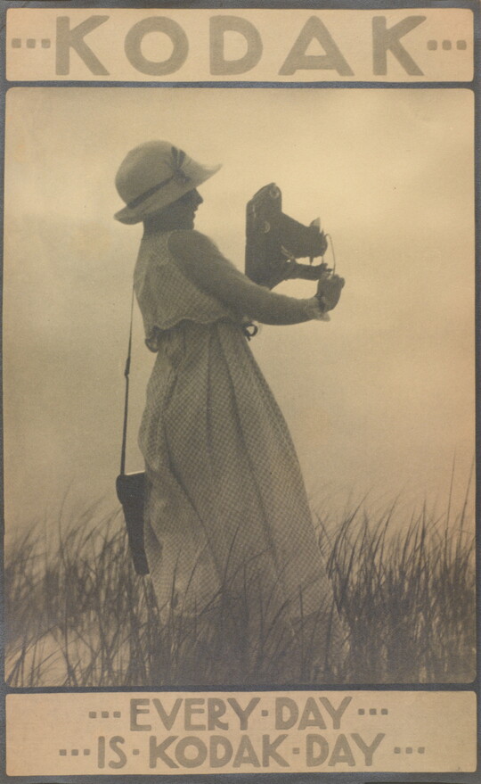 A sepia-toned photograph of a woman in a sunhat and long white dress standing in grass holding a large camera with text that reads, "Every day is Kodak day."