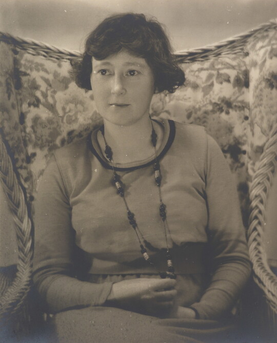 A black-and-white photograph a White woman with short dark hair wearing a long necklace and a long-sleeve top sitting in a floral, wingback chair.