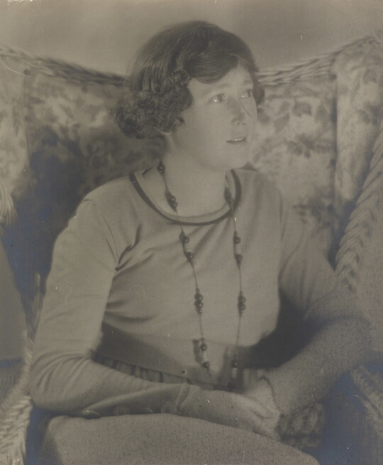 A black-and-white photograph a White woman, looking off to the side, with short dark hair wearing a long necklace and a long-sleeve top sitting in a floral, wingback chair.
