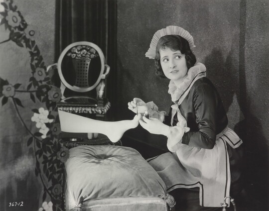 A black-and-white photograph of a White maid giving a pedicure to the foot of a woman who is hidden behind a screen.