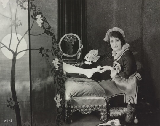 A black-and-white photograph of a White maid giving a pedicure to the foot of a woman who is hidden behind a screen.