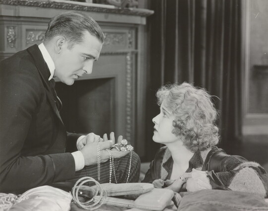 A black-and-white photograph of a White man presenting a White woman with jewelry as she gazes up at him looking worried.