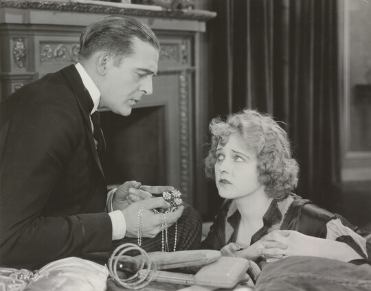 A black-and-white photograph of a White man presenting a White woman with jewelry as she gazes up at him looking worried.