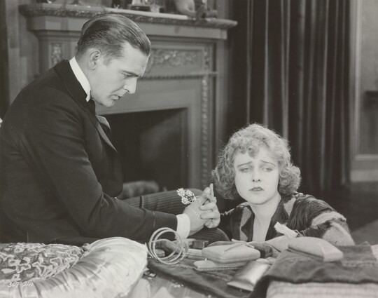 A black-and-white photograph of a White man presenting a White woman with jewelry as she sadly gazes into the distance.