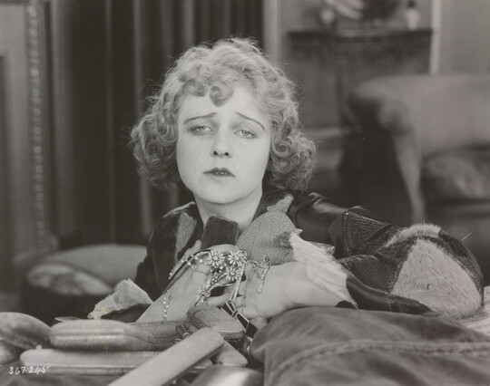 A black-and-white photograph of a White woman holding jewelry to her chest as she sadly gazes into the distance.
