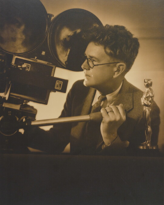 A sepia-toned photograph of a White man looking into a large movie camera and an Oscar statuette on a table next to him.