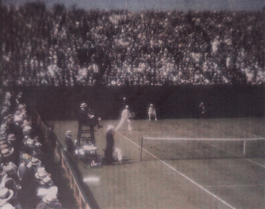 A faded color photograph of spectators in crowded stands watching a tennis match.