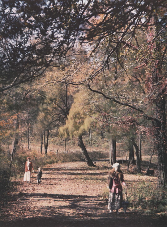 A color photograph of two adults and one child walking on a dirt road under autumn-colored trees.