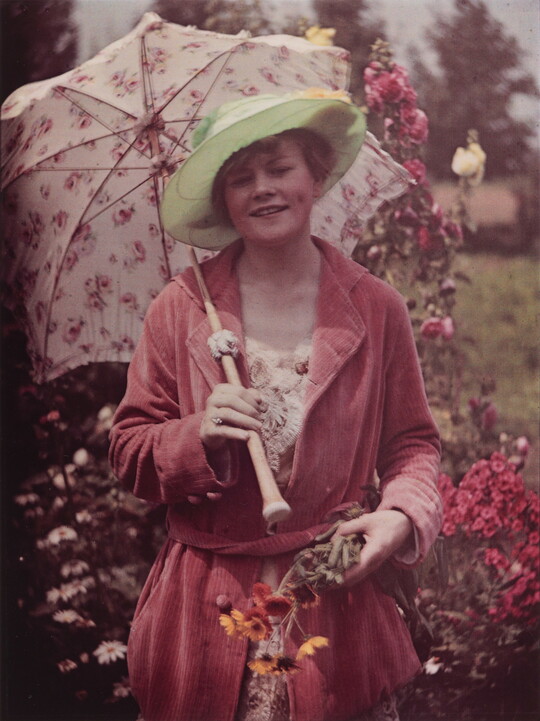 A color photograph of a smiling White woman, standing in a garden, wearing a green wide brim hat, a pink robe, holding a parasol in one hand and a bunch of flowers in the other.