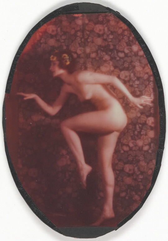 A faded color oval-shaped photograph of a nude, fair-skinned women posed as if in the middle of a dance, with one leg bent, arms out, and yellow flowers in her hair.
