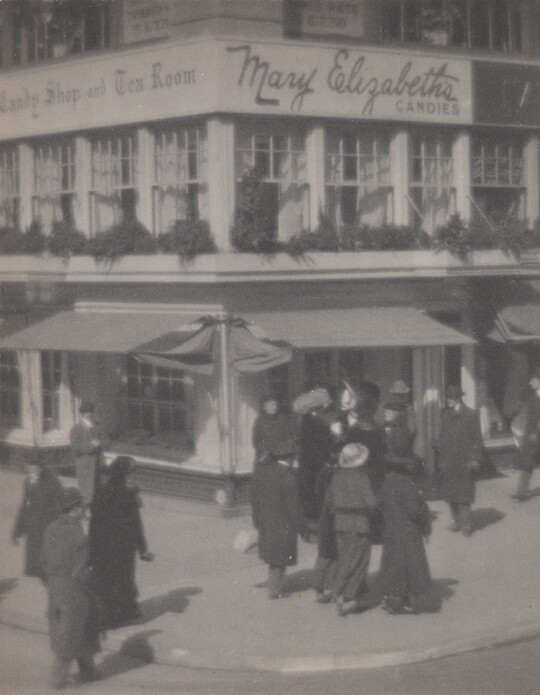 A black-and-white photograph of people walking along a street corner in front of a multi-story building with awnings at street level.