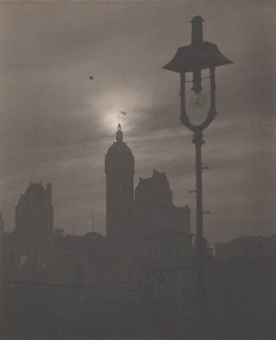 A black-and-white photograph of the silhouettes of several buildings and a gas streetlight.