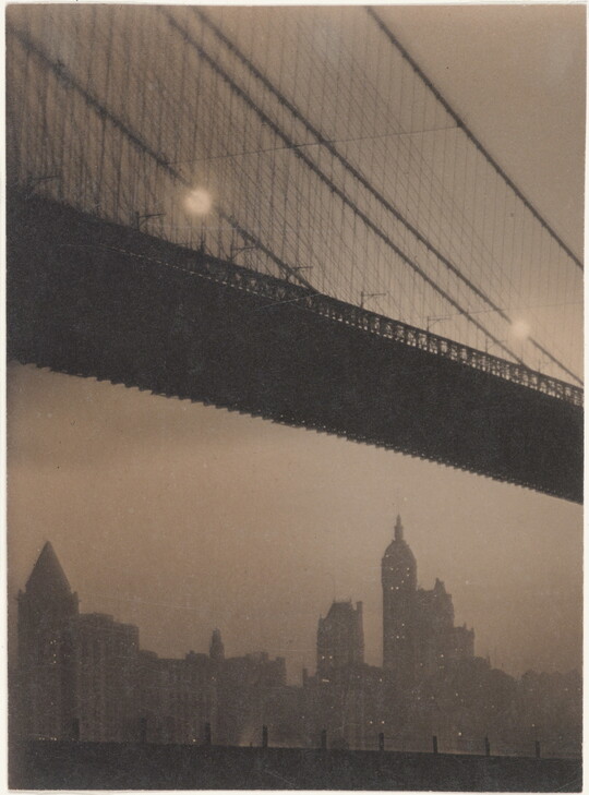A black-and-white photograph of a suspension bridge seen from below and a city skyline in the distance.