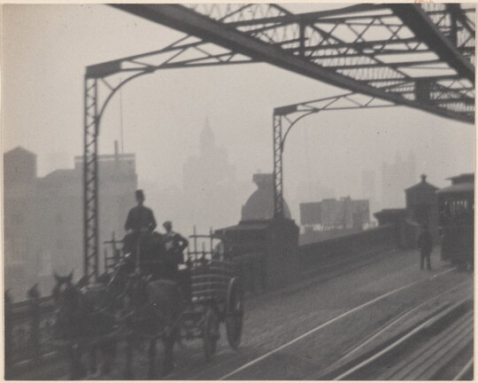 A black-and-white photograph of a horse-drawn carriage crossing a bridge next to streetcar rails as haze obscures buildings in the background.