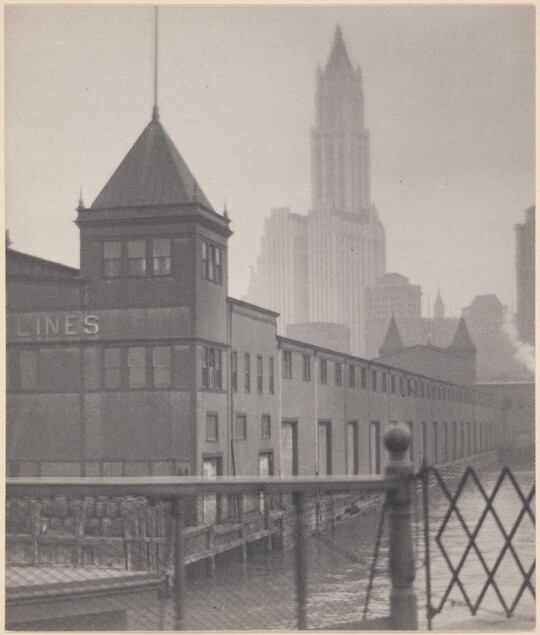 A black-and-white photograph of a low building with a tower on a canal, a gate in the foreground, and skyscrapers in the background.