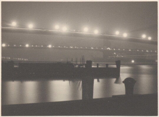 A black-and-white photograph of illuminated bridges at night reflected in the water beneath them; several ships in the water.