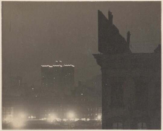 A black-and-white photograph of a building lit around the roofline and a silhouette of a closer building with a billboard on top.