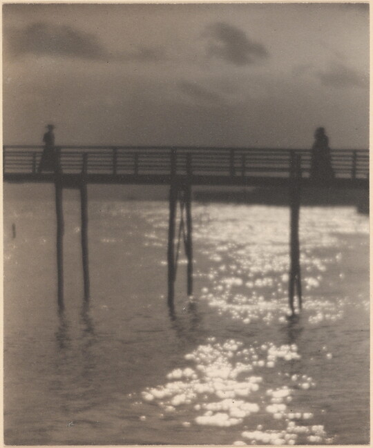 A black-and-white photograph of people walking toward each other on a pier with light reflected over the water underneath.