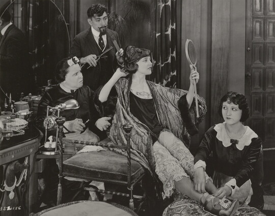 A black-and-white photograph of an elaborately-dressed White woman seated on a bench, gazing into a hand mirror, surrounded by people who are helping her dress.