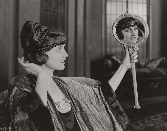 A black-and-white photograph of an elaborately dressed White woman holding up a mirror and gazing into it, her gaze in the mirror meeting the viewer's eye.