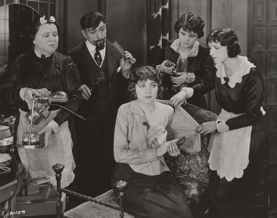 A black-and-white photograph of a White woman seated on a bench surrounded by people who are helping her undress.