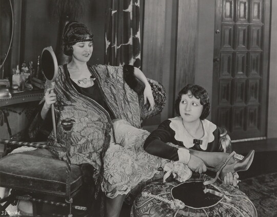 A black-and-white photograph of an elaborately-dressed White woman holding a large hand mirror, seated on a chair with one foot resting on an ottoman as a maid puts on her shoes.