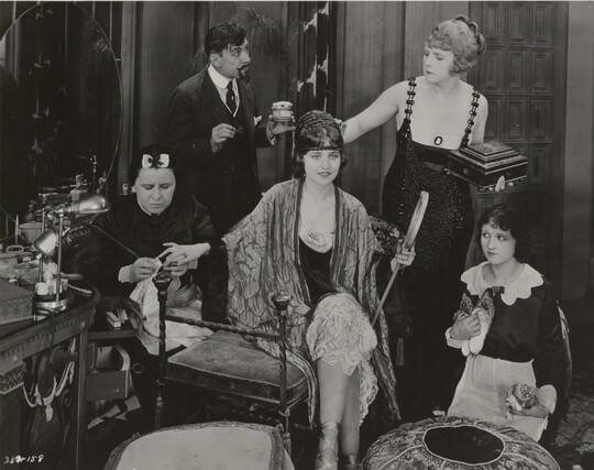 A black-and-white photograph of an elaborately-dressed White woman seated on a bench surrounded by people who are working on her hair and nails.